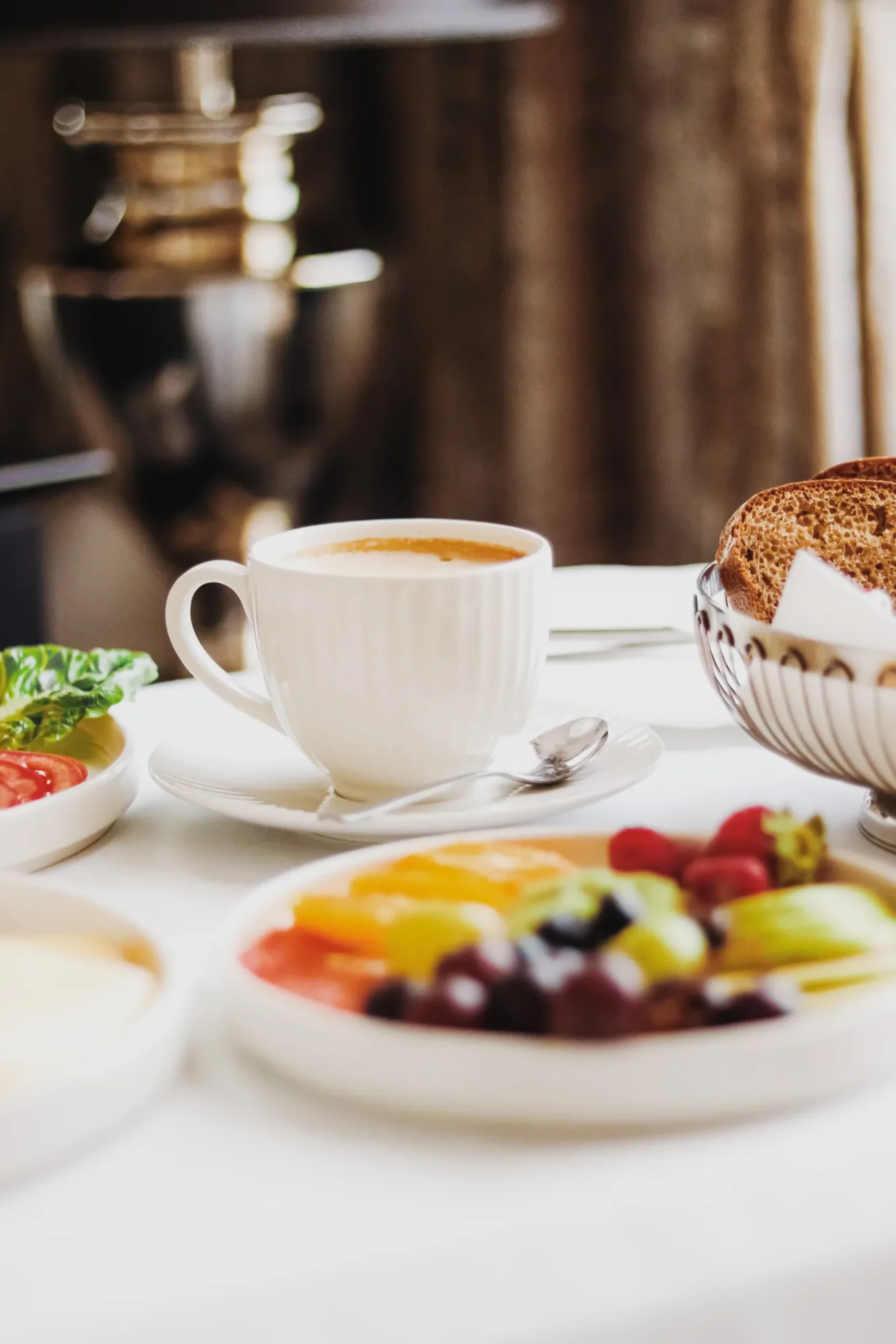 Frühstückstisch mit Kaffee, Obstsalat, Salat, Tomate und dunklem Brot in einer Silberschale auf weißer Tischdecke.