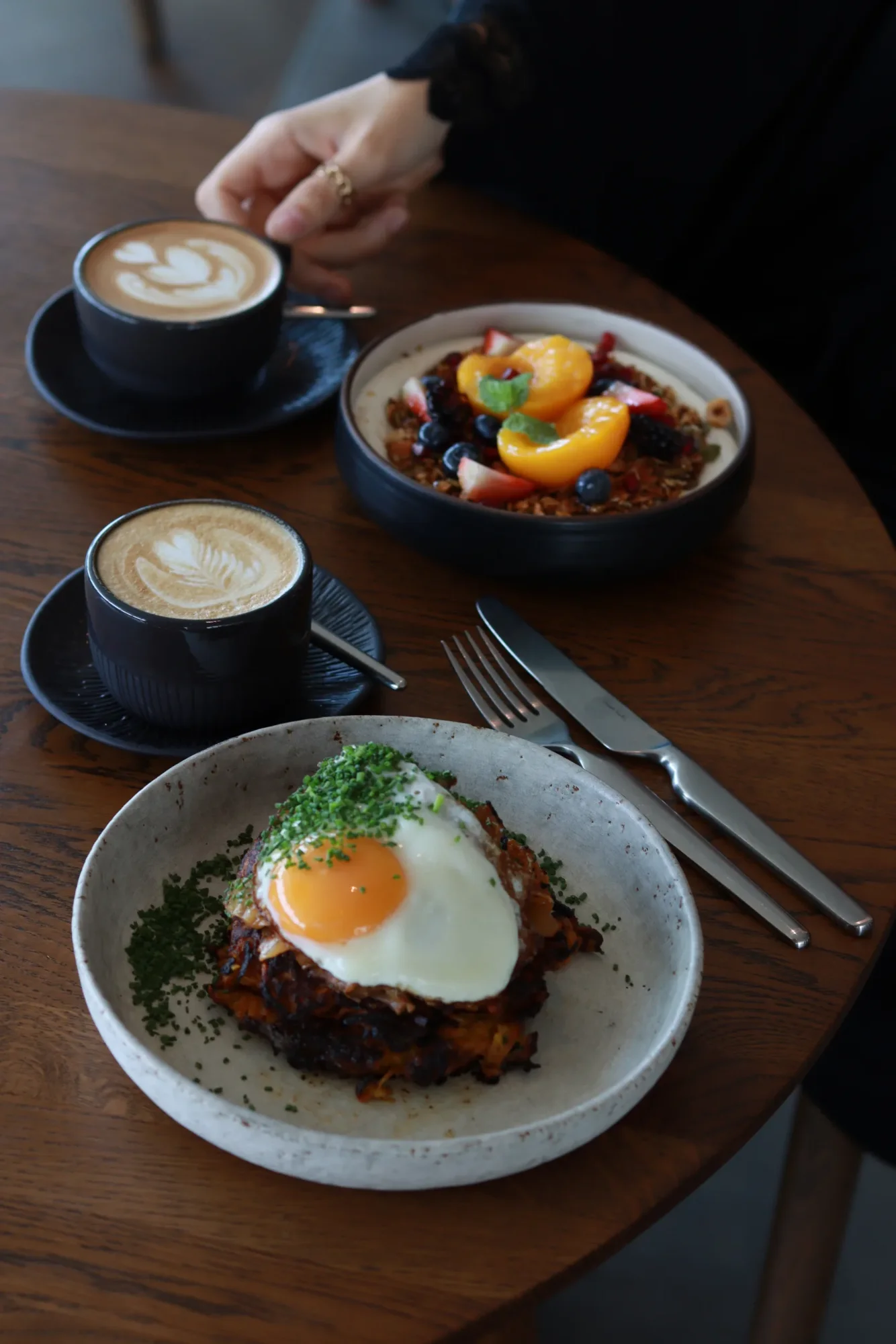 Frühstück mit zwei Kaffees, Granola mit Früchten und einem Rösti mit Spiegelei und Schnittlauch auf einem Holztisch.