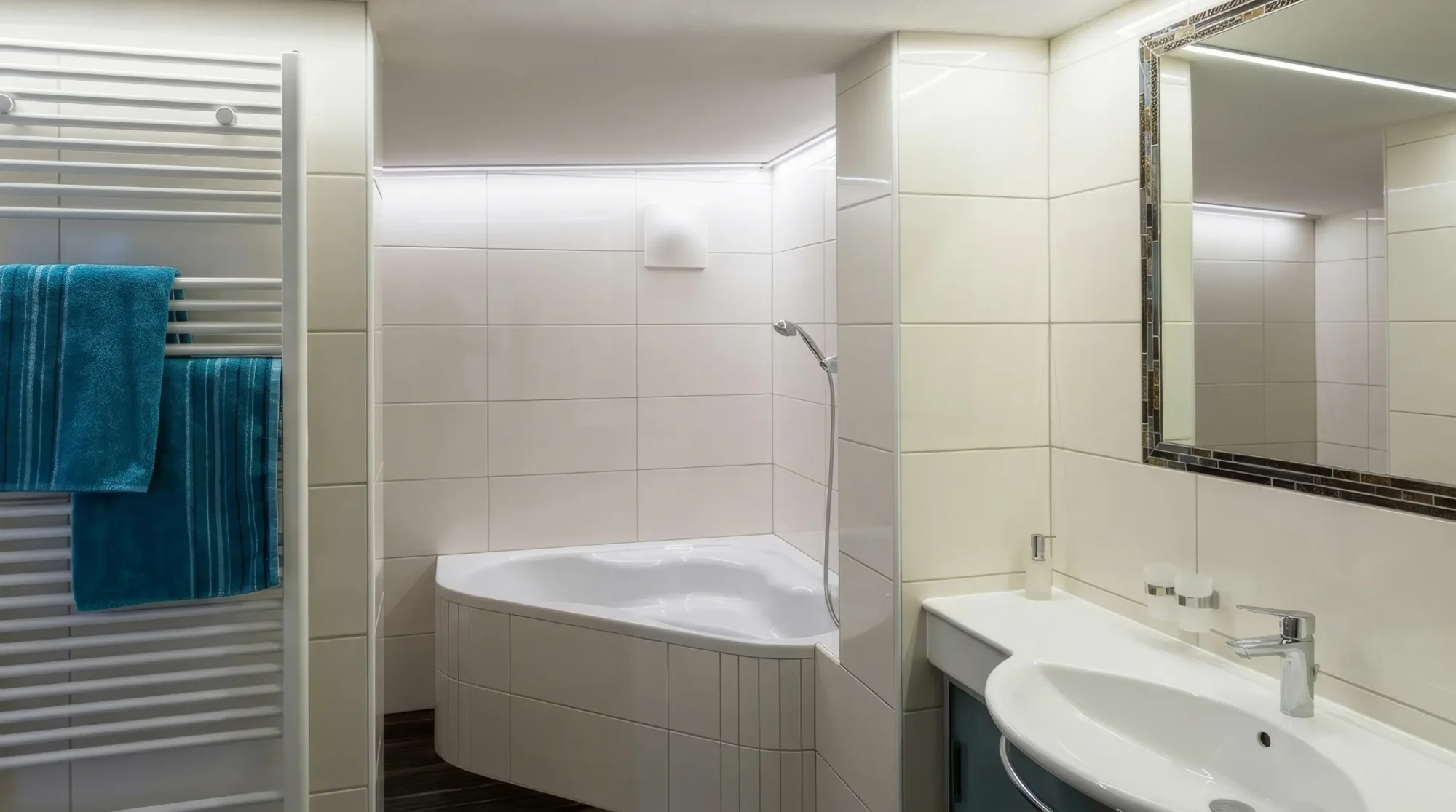Modernes Badezimmer mit weißen Fliesen, Eckbadewanne, Waschbecken, Spiegel und Handtuchheizkörper mit blauen Handtüchern.