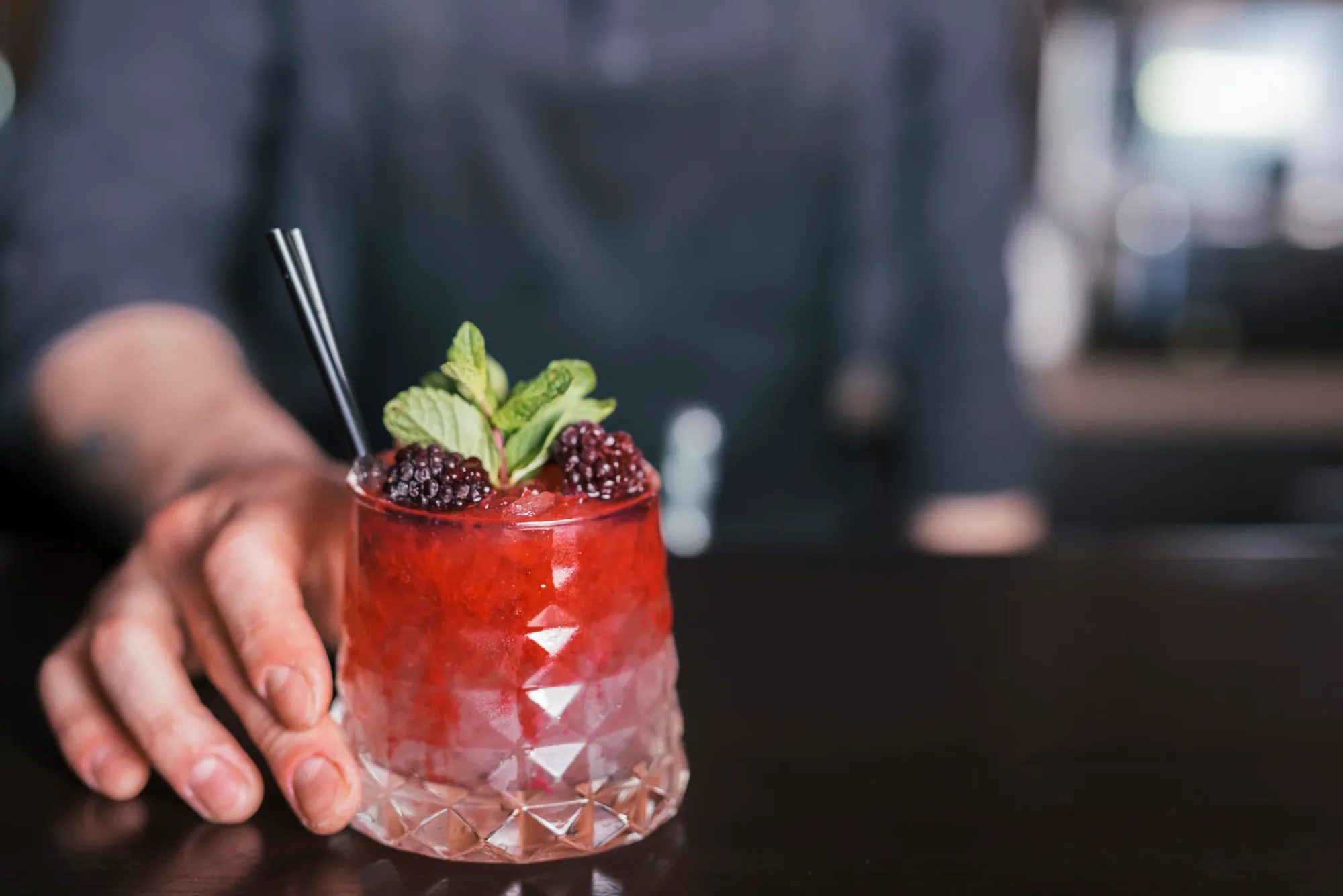 Rotes Cocktailglas mit Brombeeren, Minze und Strohhalm, serviert von einer Hand auf dunkler Theke.