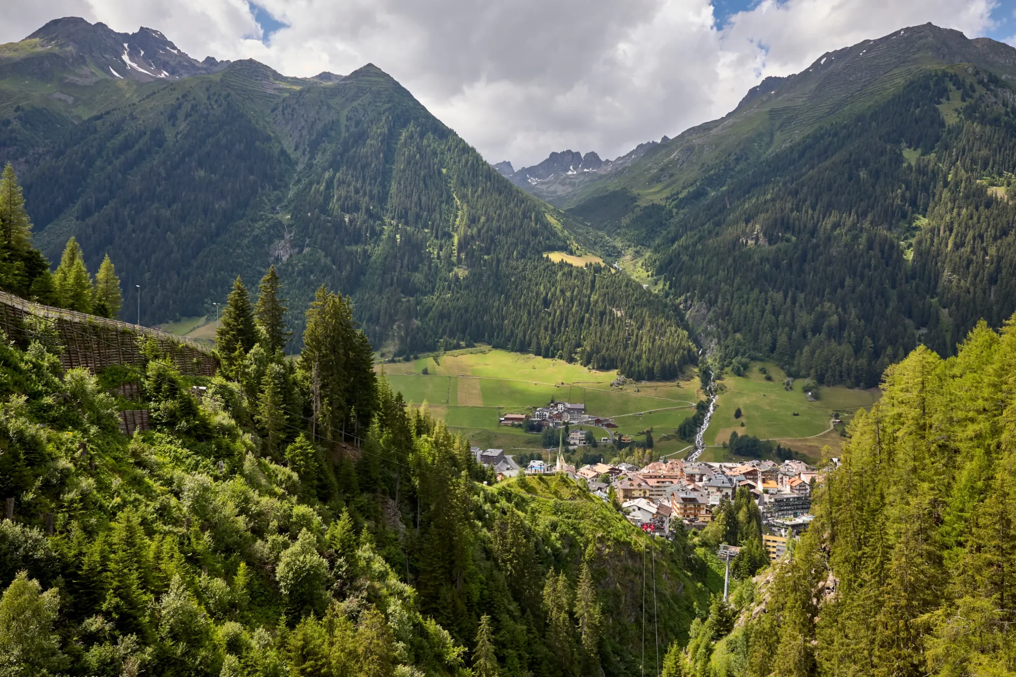 An alpine village in a green valley, surrounded by densely wooded mountains beneath a cloudy sky.
