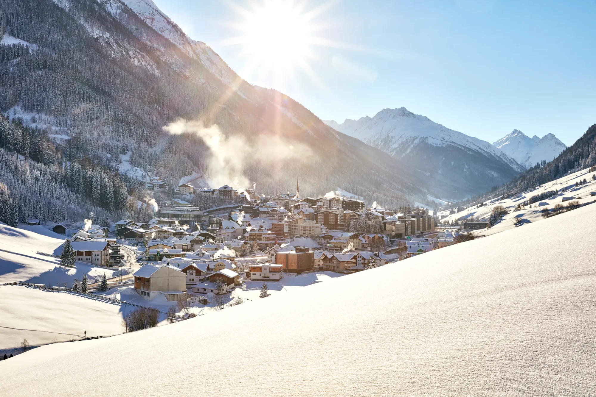 A sunny winter’s day over a snow-covered village in a deep valley, surrounded by snow-capped mountains and woods.