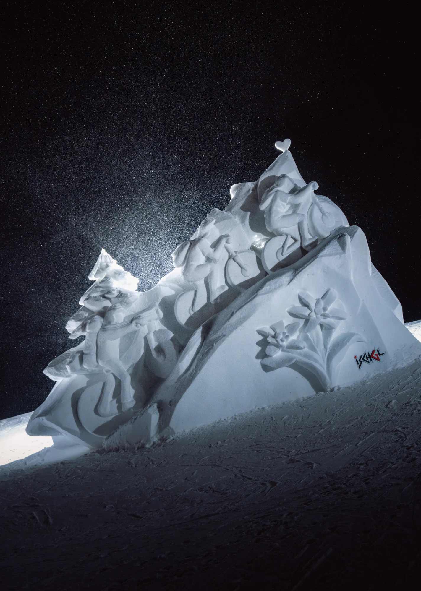 An illuminated snow sculpture featuring cyclists, a heart and edelweiss against a dark sky with glistening snow.