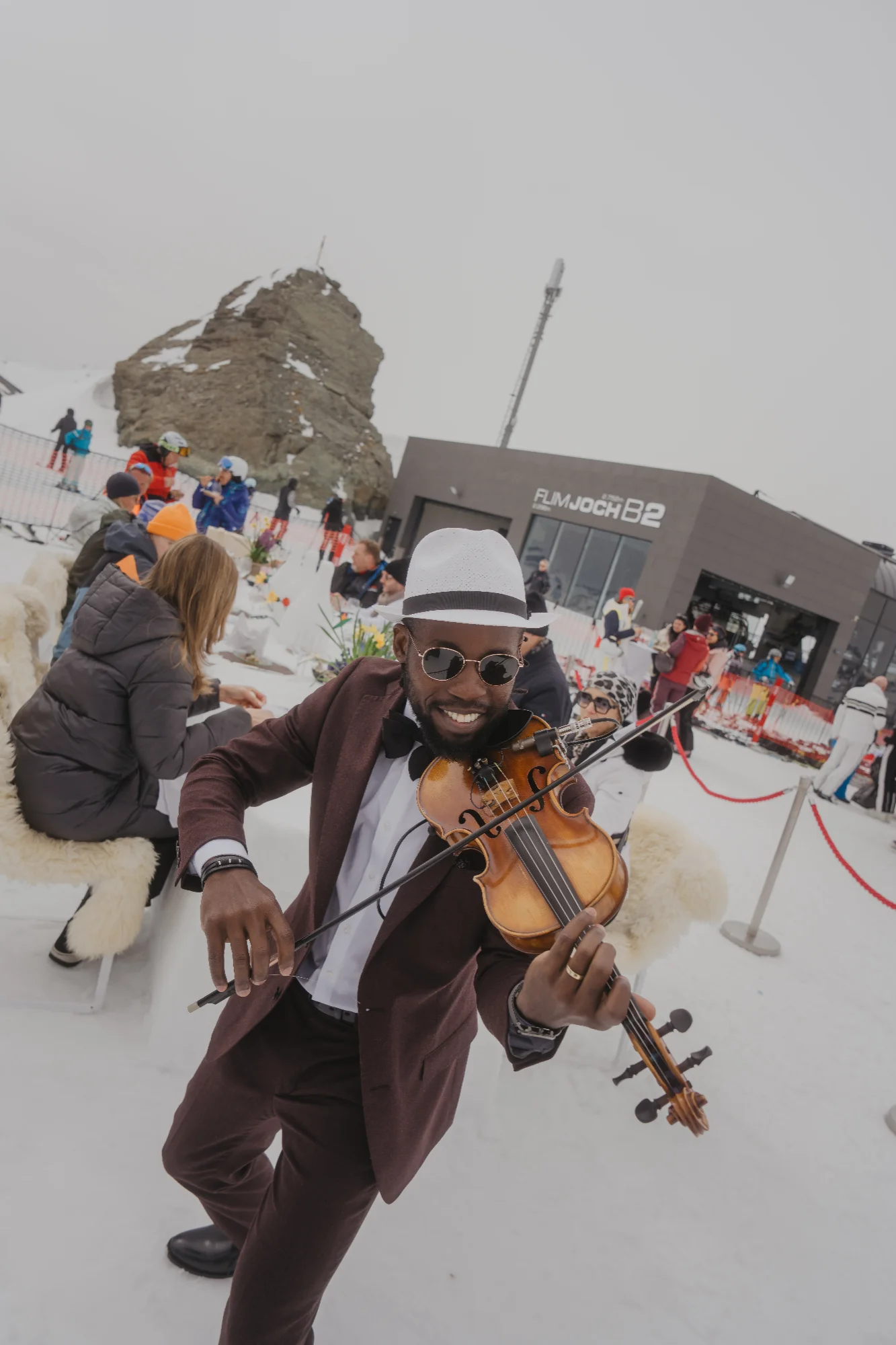 A man in a suit, hat and sunglasses is playing the violin on a snow-covered mountain, surrounded by skiers and a mountain restaurant.