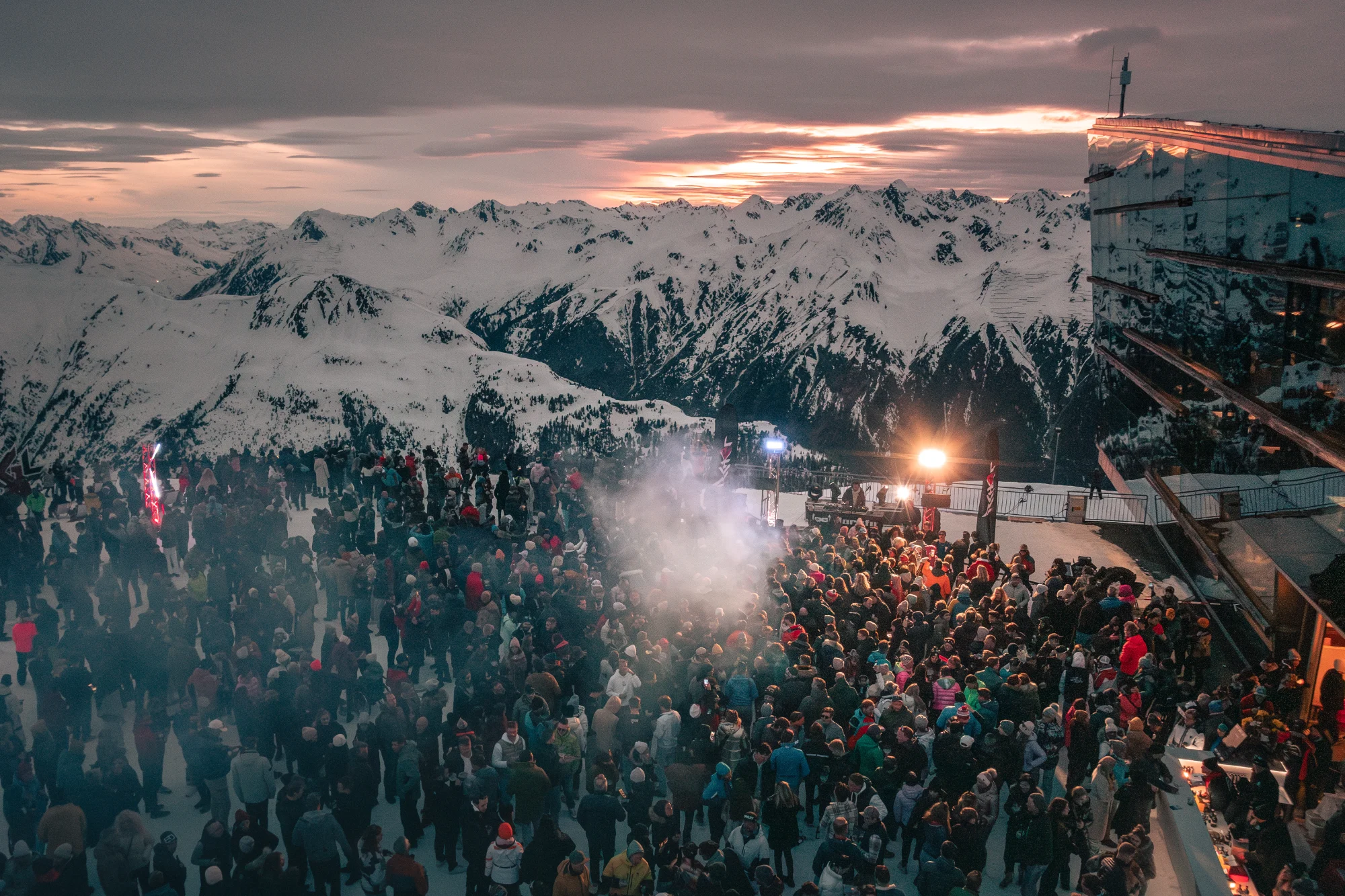 A winter party on a snow-covered mountain with a large crowd, lights and smoke, set against a stunning mountain backdrop at sunset.