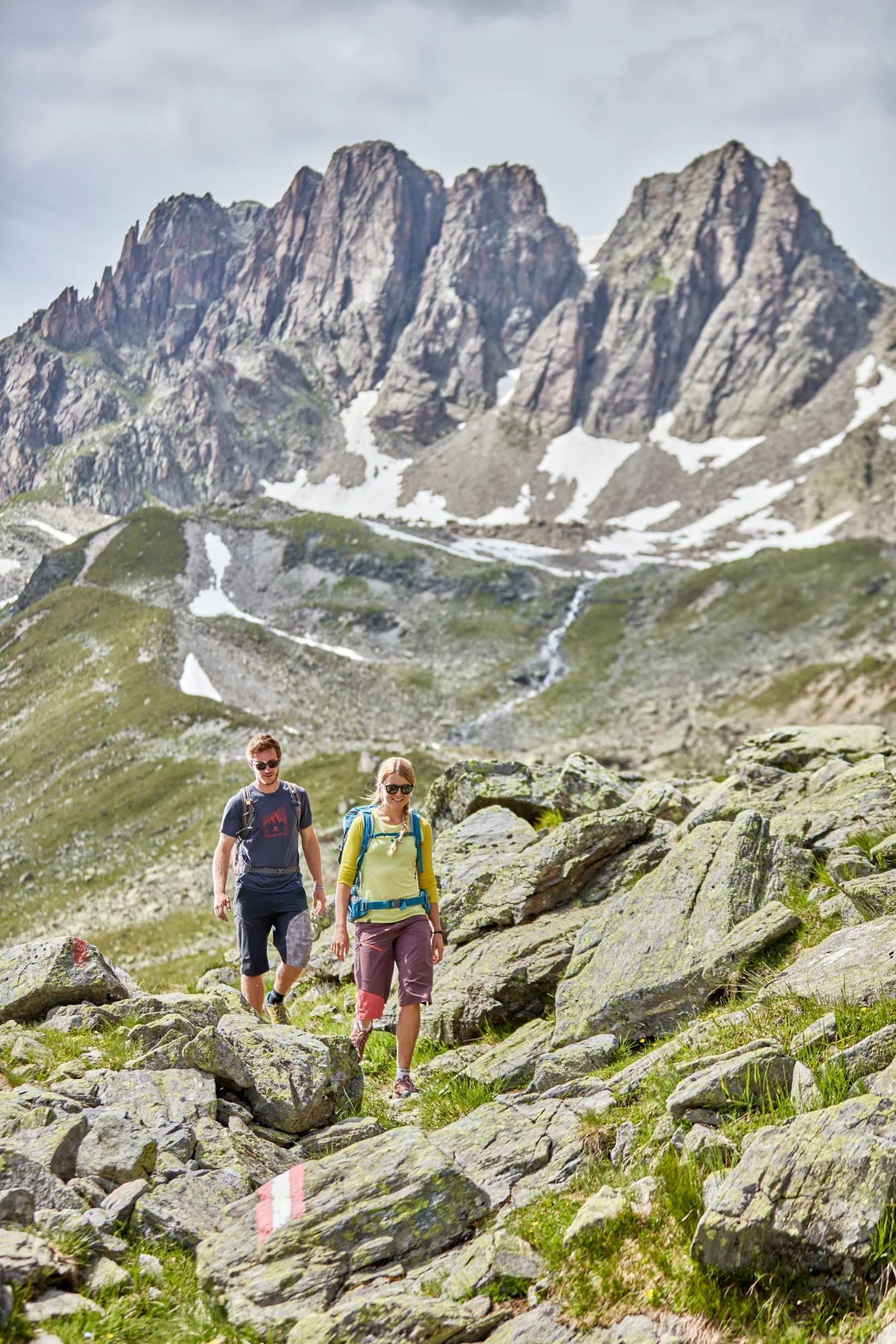 Two walkers with rucksacks on a rocky mountain path beneath high, snow-capped peaks and a cloudy sky.