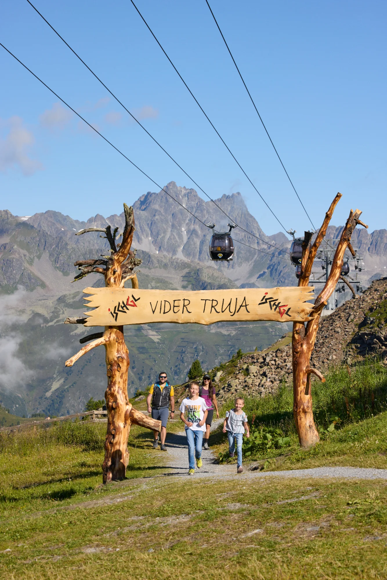A family goes hiking beneath the wooden sign ‘Vider Truja’, with the cable car and the Alpine mountain scenery in the background, in Ischgl.