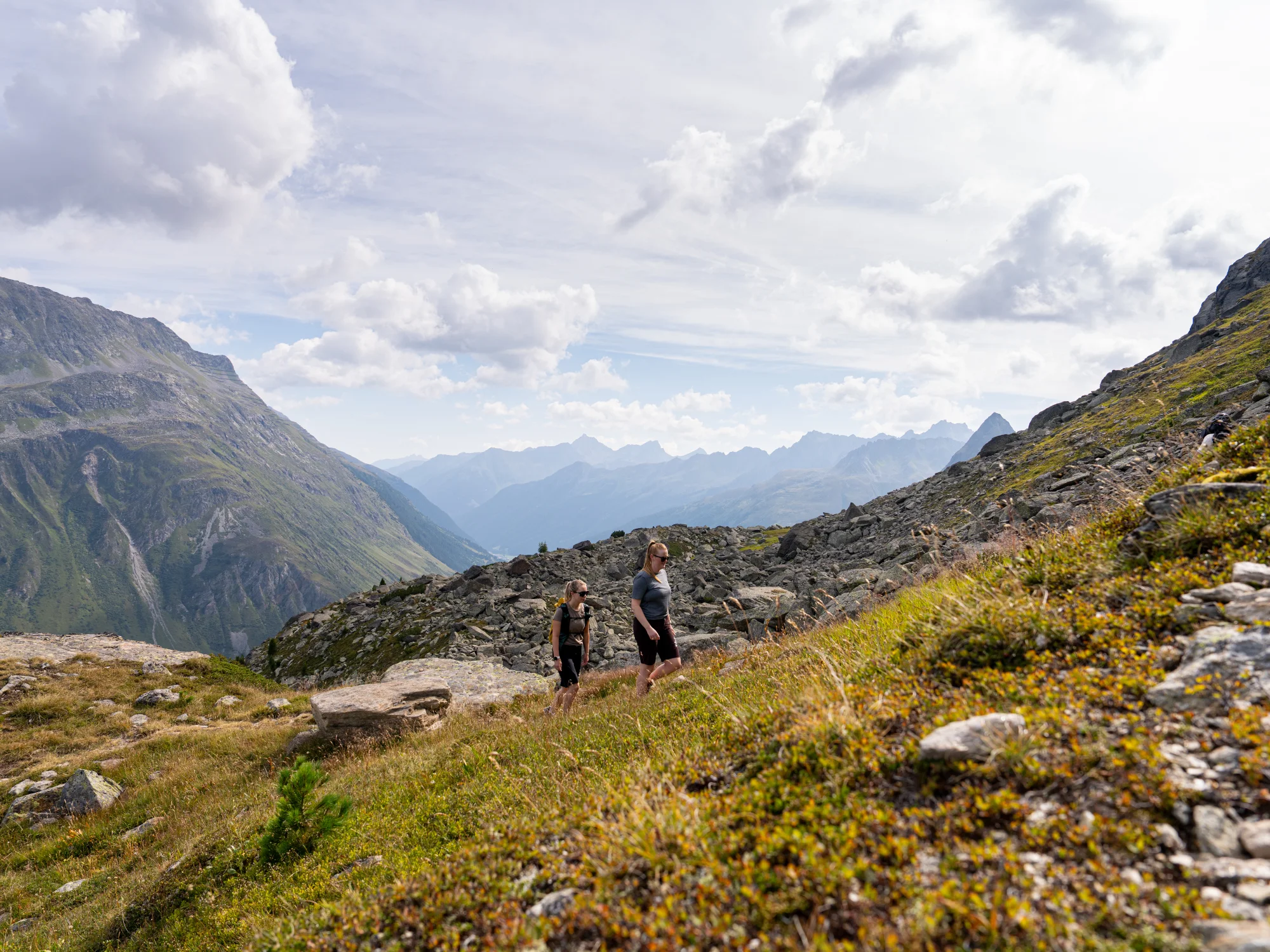 Two women are hiking up a grassy mountainside, with rocky peaks and a wide valley stretching out below them under a cloudy sky.