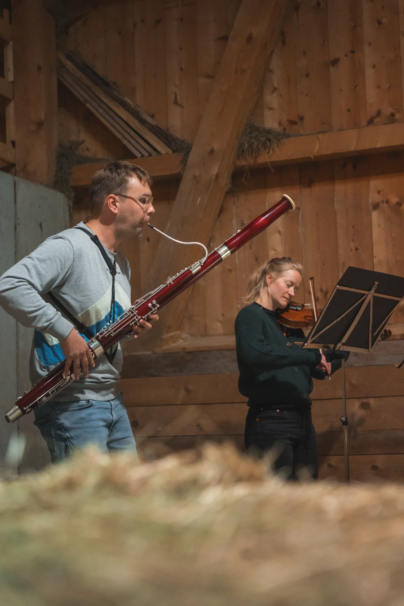 A man plays the bassoon and a woman plays the violin in a rustic wooden room filled with hay.
