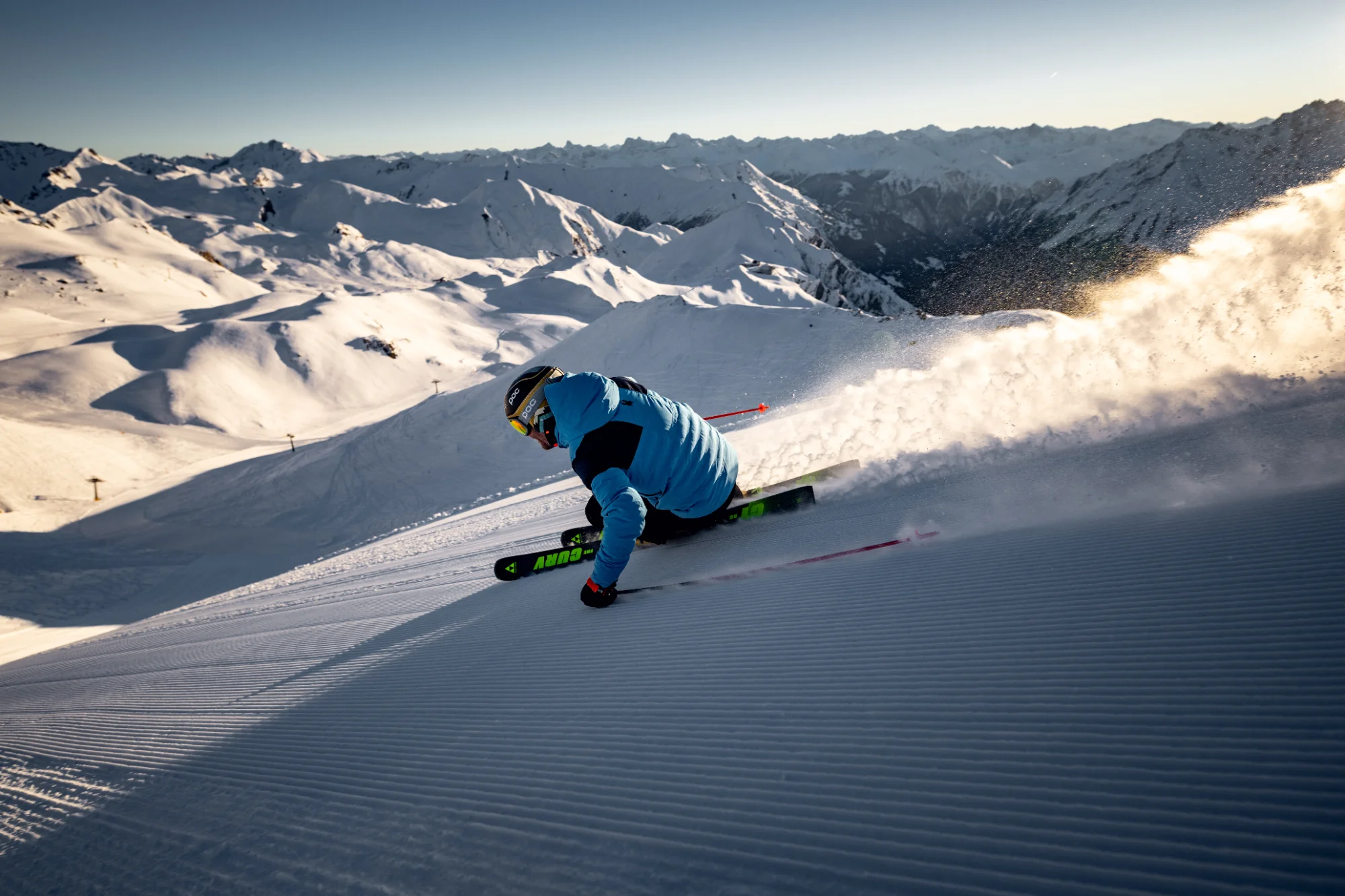 Skifahrer in blauer Jacke carvt auf frisch präparierter Piste im Sonnenschein, umgeben von schneebedeckten Bergen.