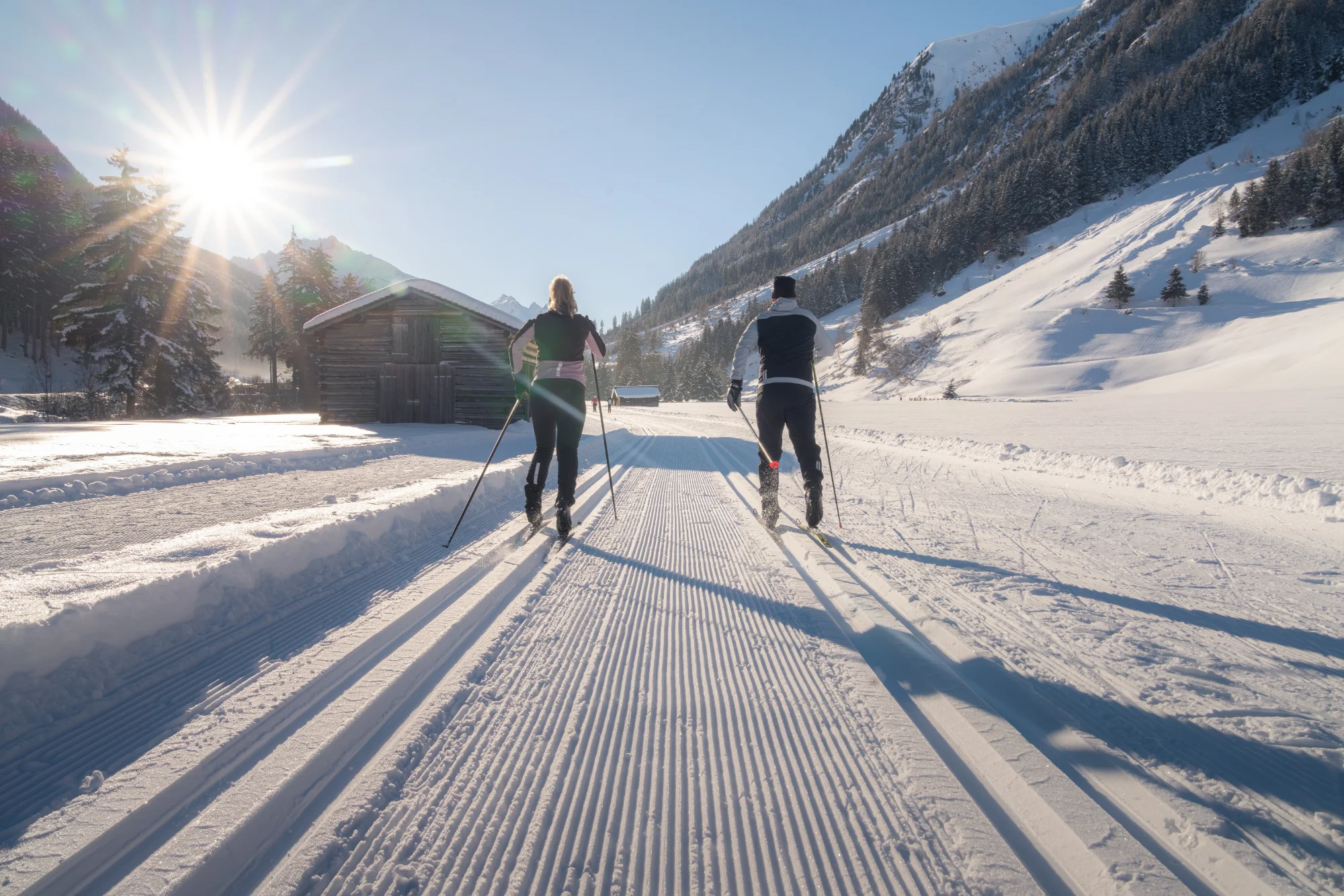 Zwei Skilangläufer auf einer präparierten Loipe im winterlichen Hochtal mit Holzhütten und verschneiten Bäumen.