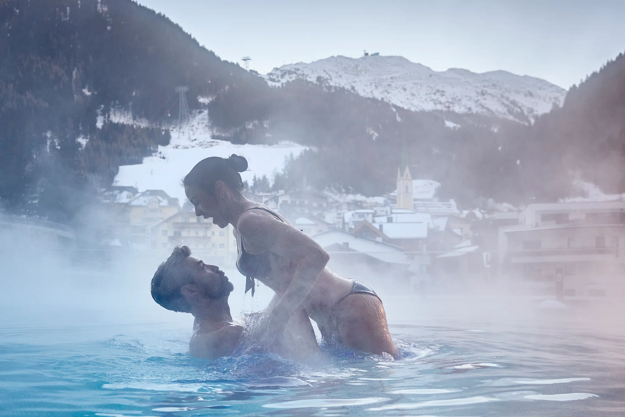 Ein Paar in einem dampfenden Pool vor einer verschneiten Bergkulisse mit Häusern.