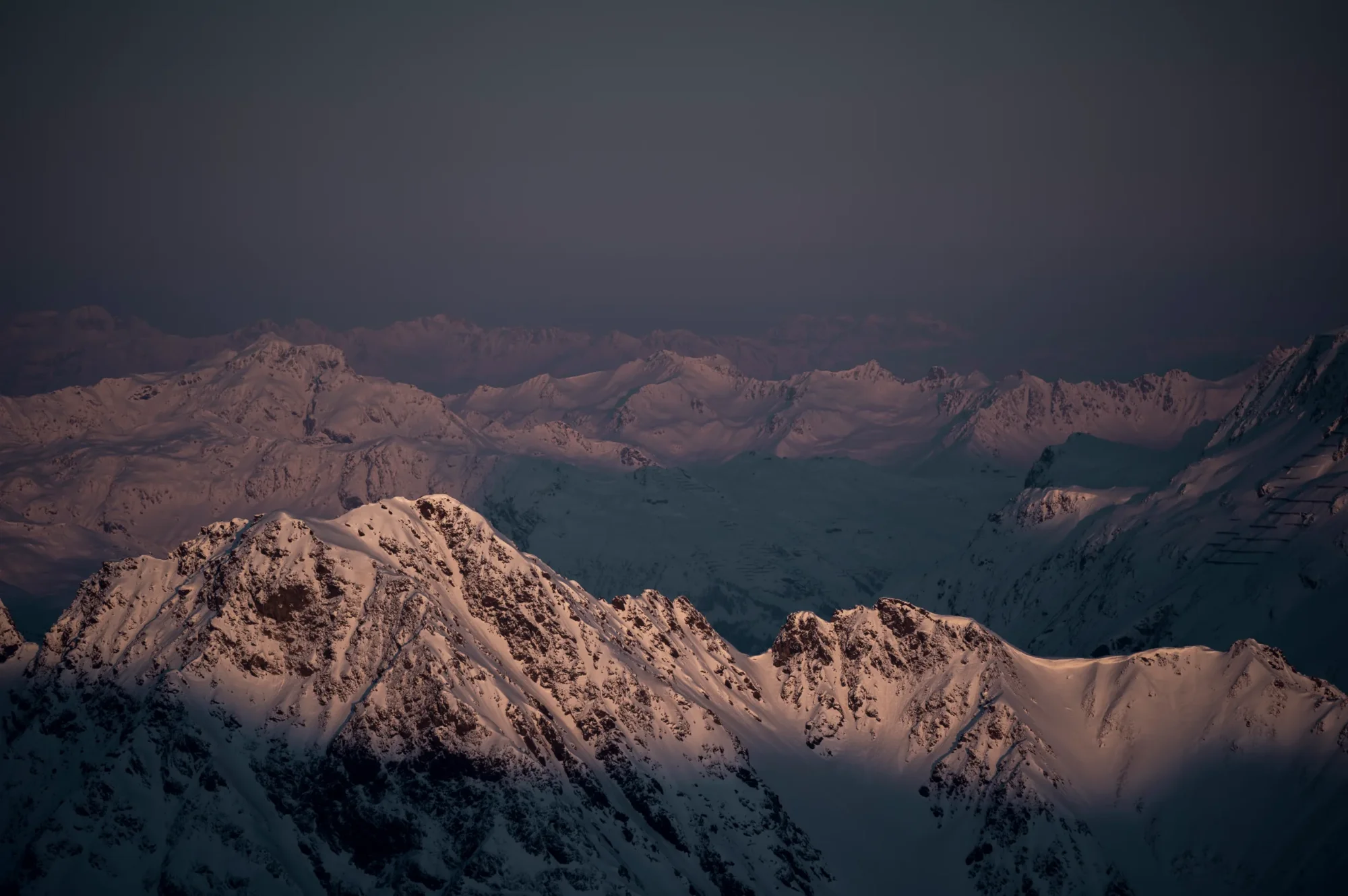Verschneite Berggipfel bei Sonnenuntergang, warmes Licht auf den Vordergrundbergen, dunkler Himmel, Bergblick.
