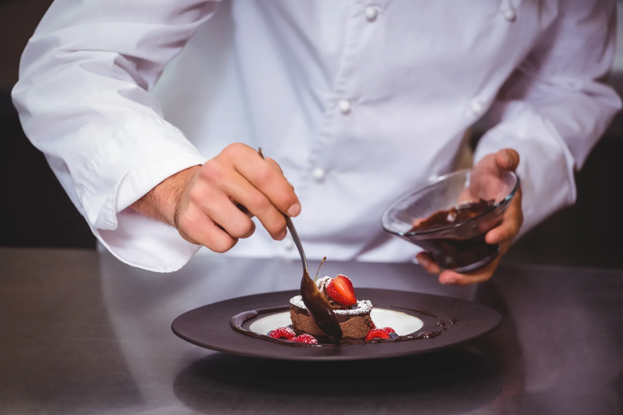Koch dekoriert Schokoladendessert mit Schokoladensauce, Erdbeere und Himbeeren auf dunklem Teller.