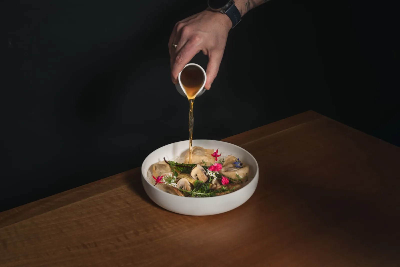 A hand pours steaming broth over a beautifully presented dish of dumplings, mushrooms and edible flowers in a white bowl.