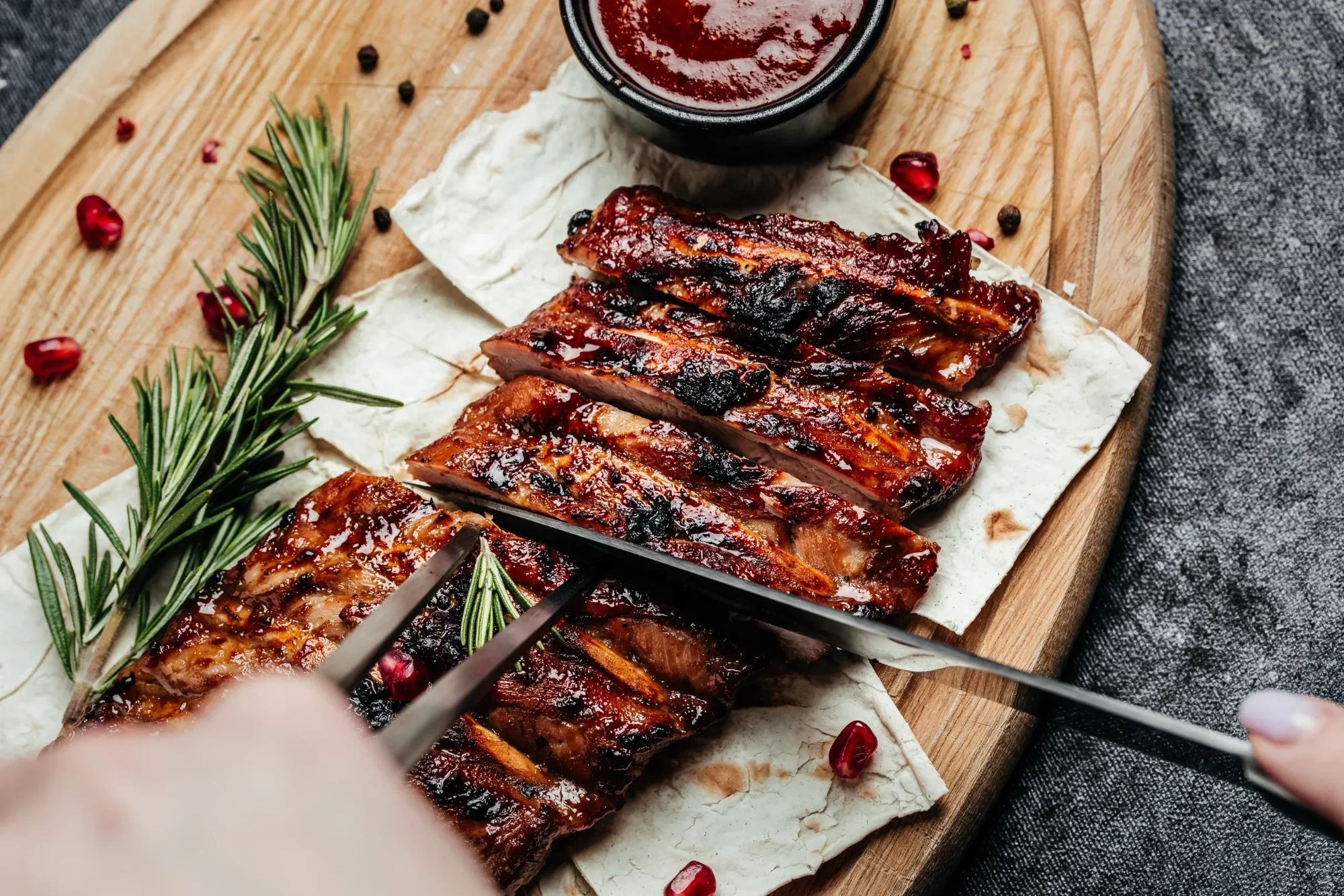 Glänzende, glasierte BBQ-Rippchen auf Fladenbrot mit Rosmarin und Granatapfelkernen werden auf Holzbrett geschnitten.