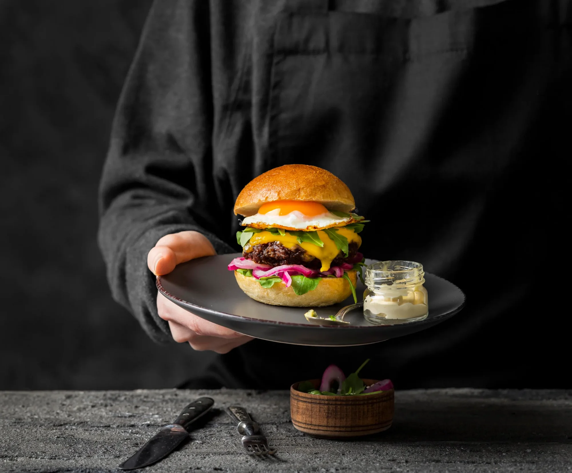 Hand reicht Teller mit Burger: gebratenes Ei, Käse, rote Zwiebeln, Grünzeug; daneben Mayonnaise im Glas.