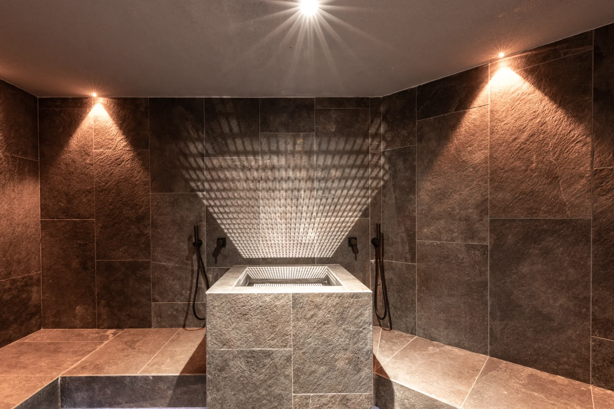 A modern hammam with dark stone tiles, a central pool and indirect lighting that casts patterns onto the wall.