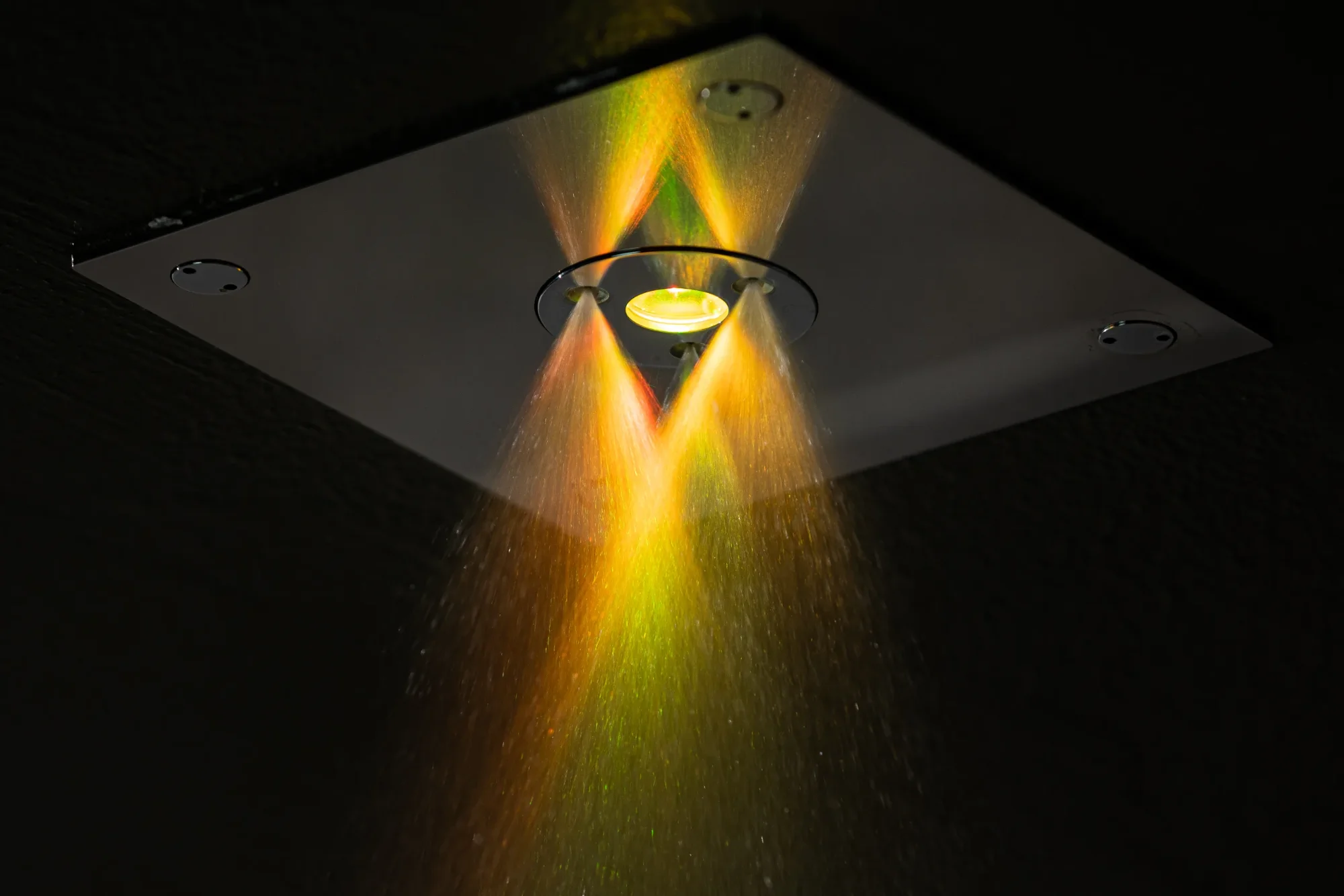 Ceiling shower with coloured water jets in red, orange and green, fitted to a dark ceiling.