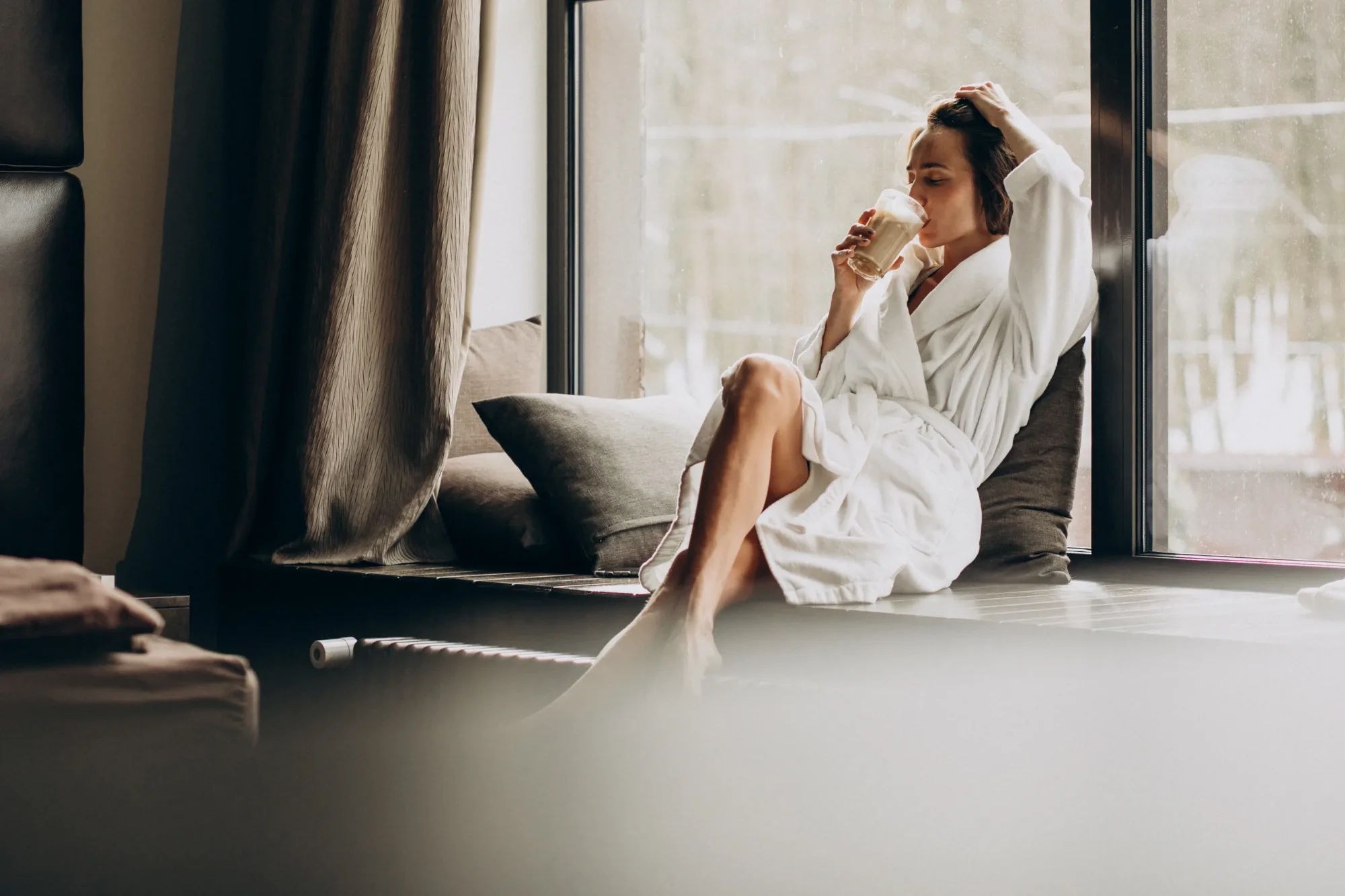 A woman in a white bathrobe is sitting by the window, drinking from a glass and relaxing in the natural light.