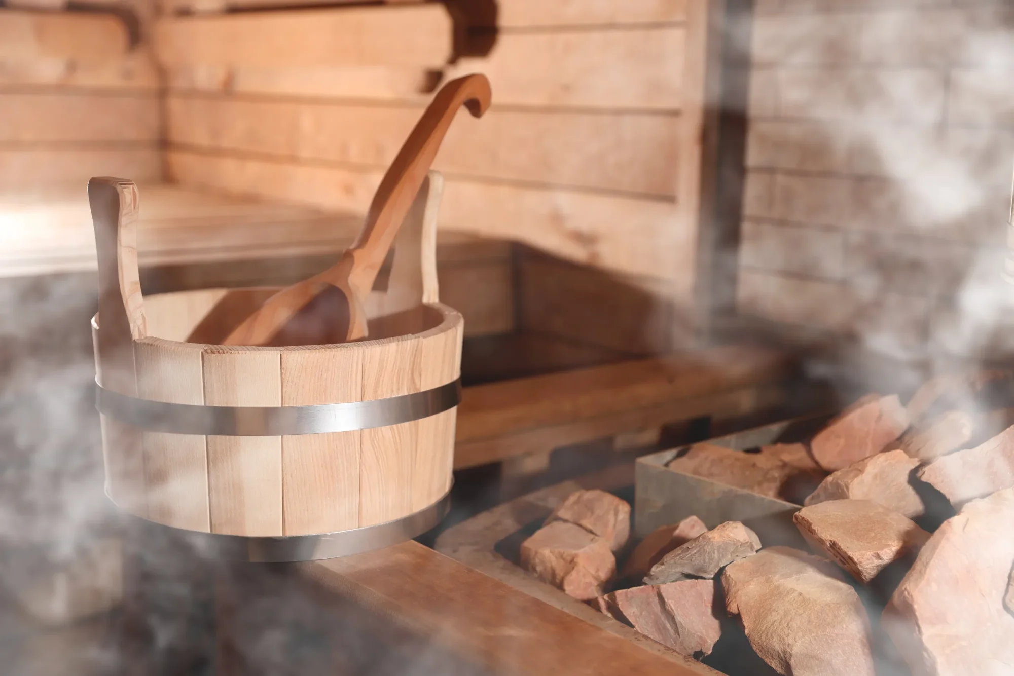 A wooden sauna bucket with a ladle and steaming sauna stones in front of a light-coloured wooden wall in a sauna.
