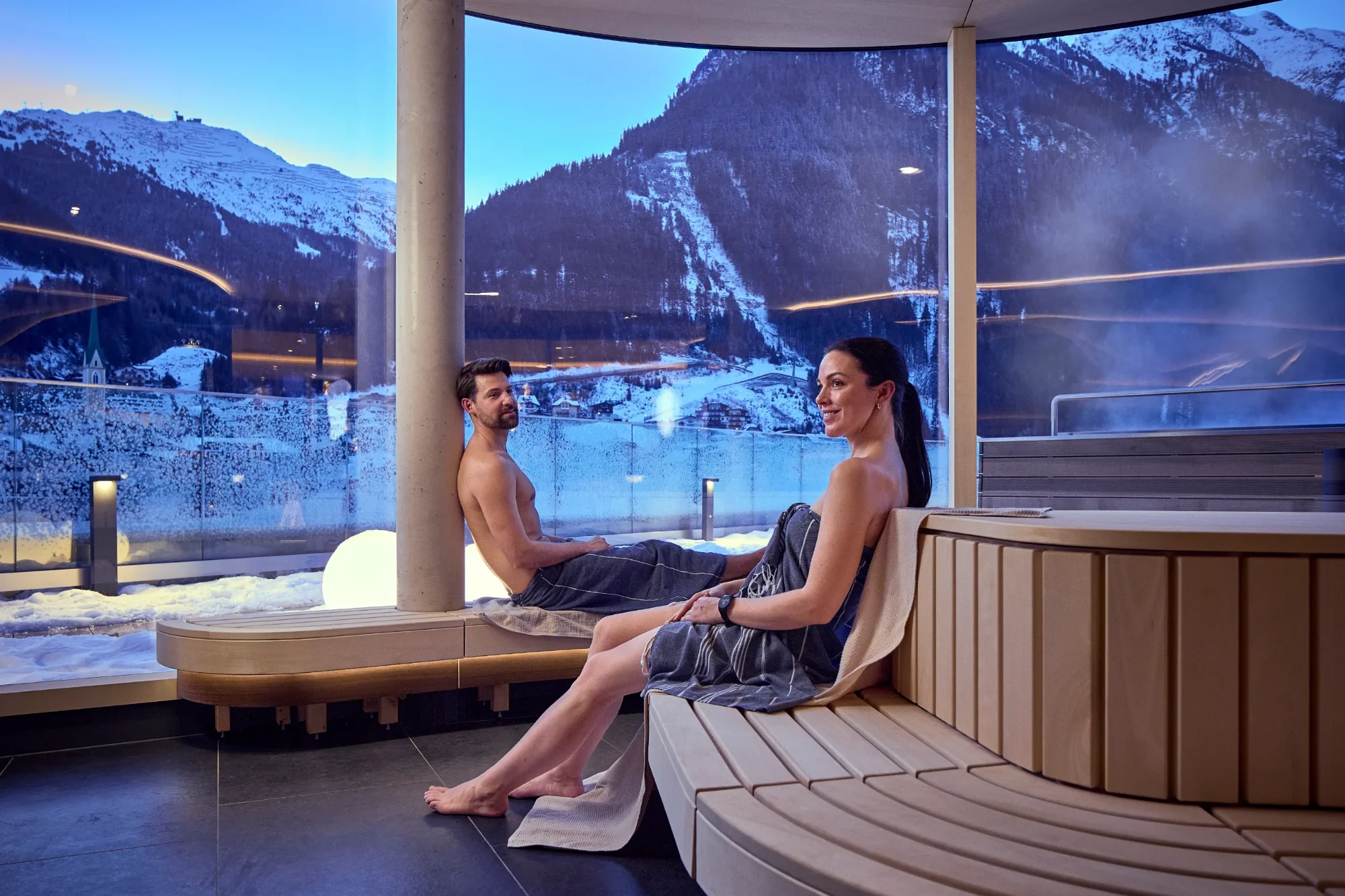 A couple at the spa, relaxing on a wooden bench in front of a panoramic window overlooking snow-capped mountains and a winter landscape.