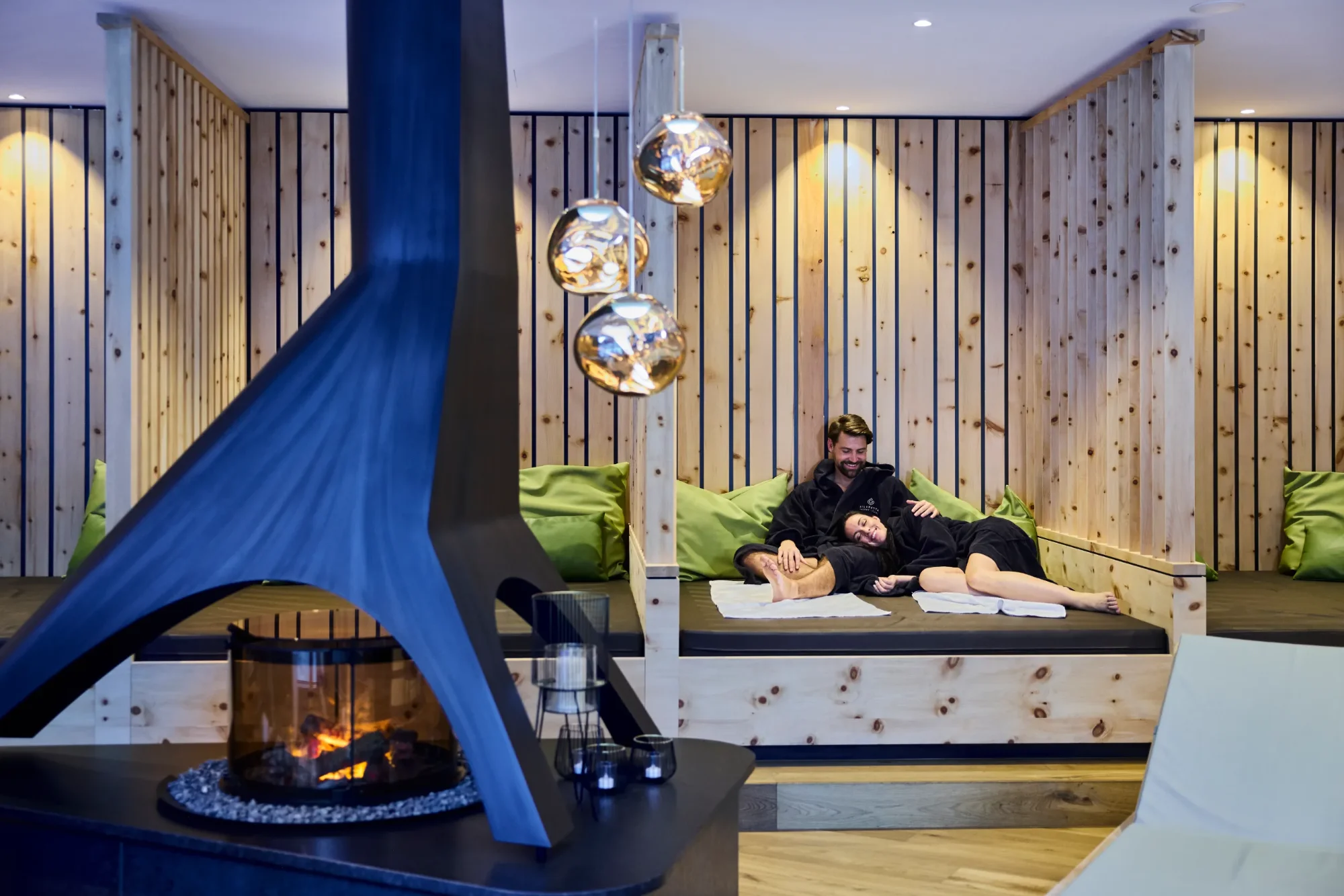 A couple relaxing in bathrobes on daybeds with wooden panels and a fireplace in a spa area.