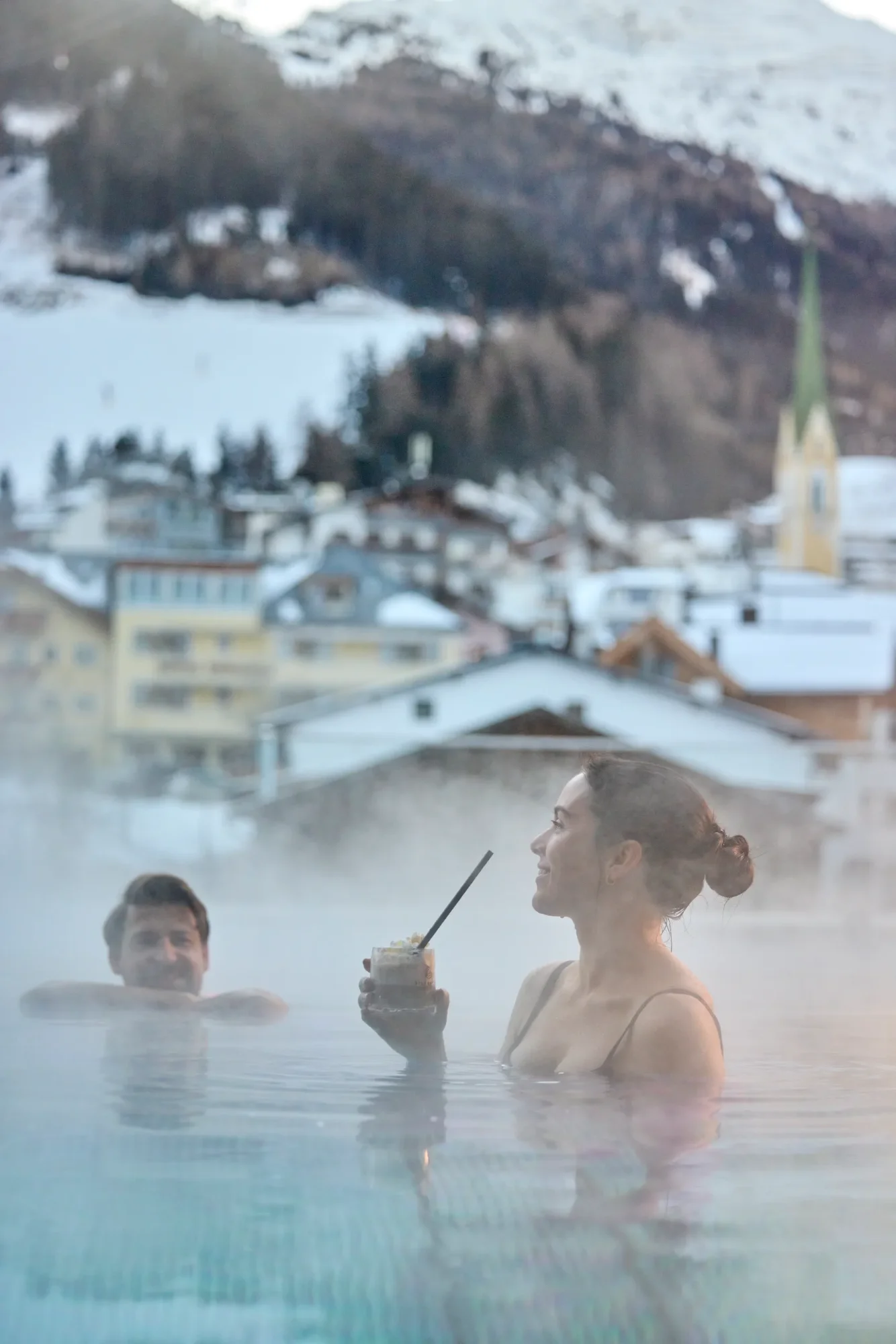 A couple in a steaming outdoor pool, sipping a drink and smiling, set against the backdrop of a snow-covered mountain village with a church. Winter holiday.