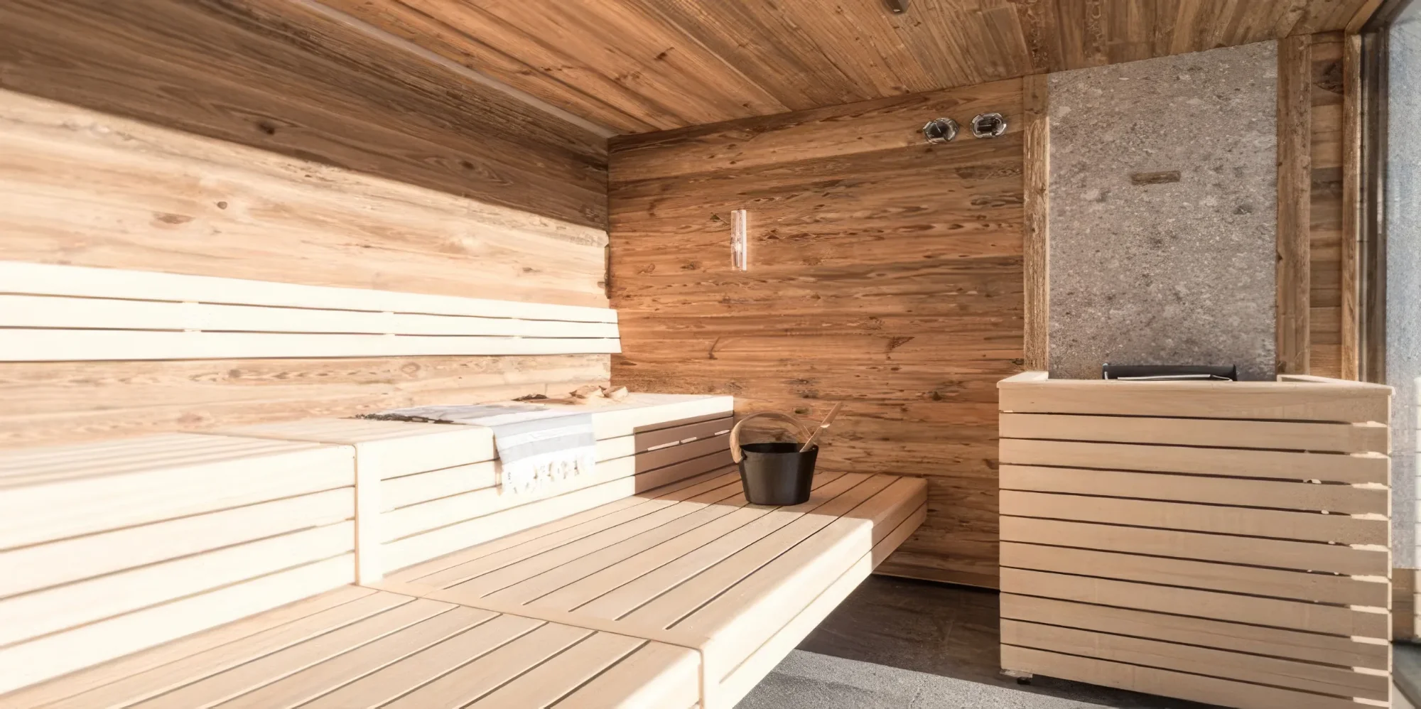 A modern sauna cubicle with light-coloured wooden benches, darker wooden walls and a stone sauna heater. A bucket, a ladle and a towel.