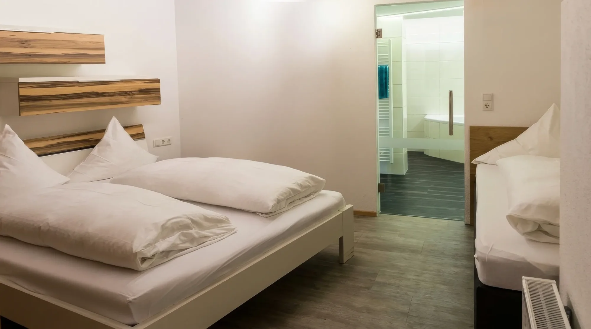Blick in ein modernes Schlafzimmer mit einem Doppelbett, einem Einzelbett und einem angrenzenden Bad mit Glasabtrennung.